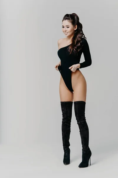 Image of satisfied brunette lady dressed in sexy black lingerie and jackboots, has slim figure and slender legs, looks gladfully down, isolated over white background. Erotic, femininity concept — 스톡 사진
