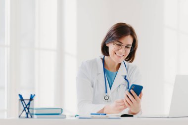 Sohbete katılan, cep telefonu tutan, mesaj yollayan, tıbbi önlük giyen, arkadaş canlısı bir ifade takınan, masaüstünde poz veren, hasta için online danışmanlık veren olumlu bir kadın doktor.