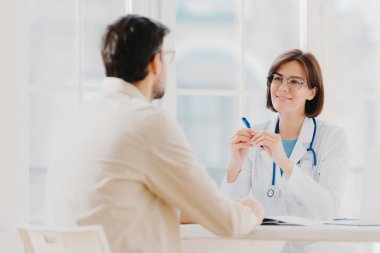 Sağlık ve tıbbi danışmanlık konsepti. Beyaz üniformalı yetenekli terapist hastayla konuşuyor, tıbbi geçmişi yazıyor, tüm semptomları dikkatle dinliyor, ciddi hastalıkları önlemeye çalışıyor.