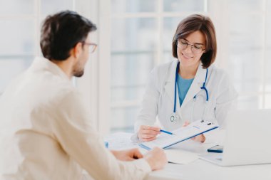 Tıbbi bir randevu. Kadın doktor erkek hastaya profesyonel tıbbi yardım yapıyor, yazılı bilgileri panoya yazıyor, destek veriyor ve iyi hizmet veriyor, masaüstü yakınlarındaki hastanede poz veriyor..