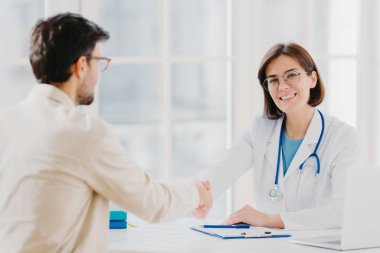 Ortaklık, yardım, güven ve tıp konsepti. Kadın doktor, iyi tedavi ve profesyonellik için müteşekkir hastayla el sıkışıyor, klinikte poz veriyor, beyaz masanın yanında tıbbi kayıtlar var.
