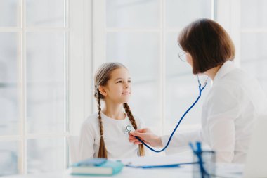 Küçük sevimli bir kız, steteskopla ciğerlerini dinleyen uzman bir doktorun muayenesini dinliyor. Sağlık kontrolüne geliyor. Çocuk sağlığı ve klinik ziyaret kavramı