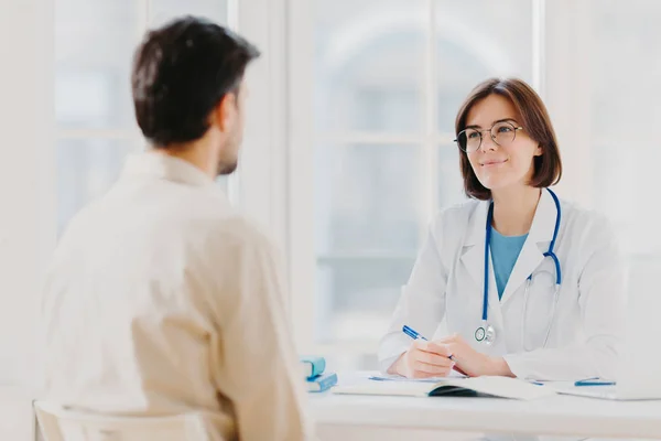 Doutor e paciente discutem algo, sentam-se à mesa na clínica. cardiologista feminino em óculos dá consulta médica de diagnóstico, conselhos para o homem como curar a doença, posar no quarto do hospital . — Fotografia de Stock