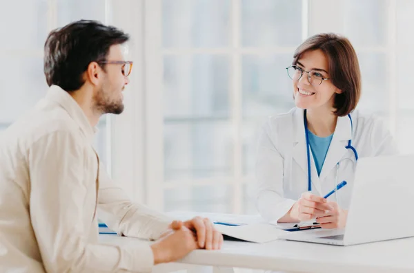 Attentive female doctor explains diagnosis to patient, gives professional consultancy, points with pen at laptop, talk about health concerns, pose at private clinic in modern office, show prescription — Stock Photo, Image