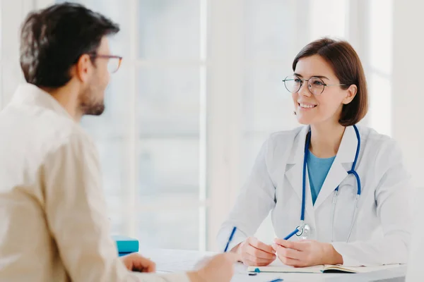 La fotografia di paziente maschile scrive tutti i dettagli fa note durante consulenza con dottore si siedono in ufficio spazioso. Il medico racconta problemi di trattamento e spiega la diagnosi ai visitatori dell'ospedale. Concetto medico — Foto Stock