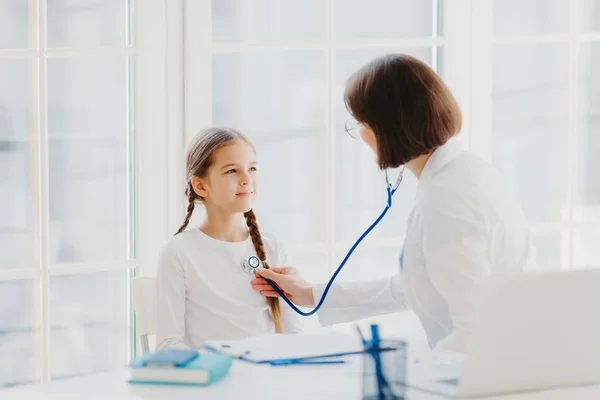 La mujer pediatra da la consulta a la muchacha pequeña, habla sobre la salud y los síntomas, escucha el corazón con el fonendoscopio, hace la prescripción, posan en la oficina del hospital. Seguro médico y atención infantil —  Fotos de Stock