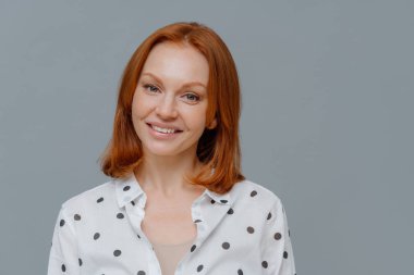 Zarif kızıl saçlı kadın kameraya nazik, dişlek bir gülümsemeyle bakıyor, sağlıklı çilli bir teni var, iyi haberler duymaktan mutlu, şık bir gömlek giyiyor, gri duvarın üzerinden izole edilmiş, metnin için kopyalama alanı var.