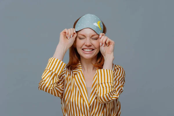 Good morning, lifestyle. Smiling freckled woman takes off blindfold, wears yellow striped pajama, awakes with smile after seeing pleasant dreams, feels relaxed and refreshed, poses over grey wall