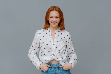 Modaya uygun pozitif bir kadın polka noktalı gömlek ve kot pantolon giyer, ellerini cebinde tutar, gri arka plana karşı kendine güvenir, işgüzar ile mutlu bir konuşma yapar. Duygular, moda konsepti