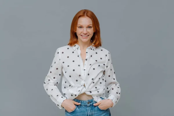 Fashionable positive woman wears polka dot shirt and jeans, keeps hands in pockets, stands self assured against grey background, has happy casual talk with interlocutor. Emotions, fashion concept