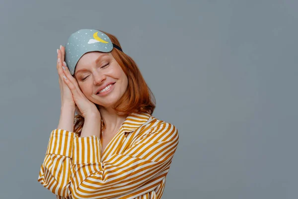 Satisfied ginger woman has nap, sees pleasant dreams, tilts head on palms, smiles gently with closed eyes, wears sleep mask, dressed in striped pajama, isolated over grey background, empty space