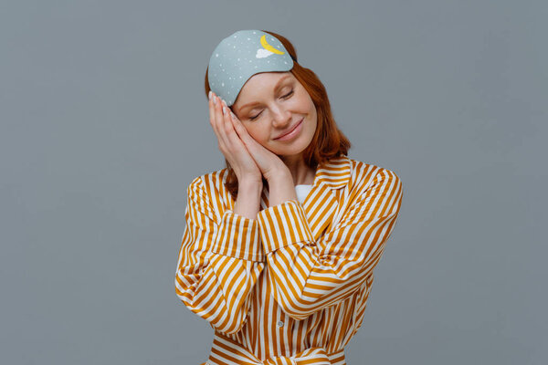 Pleased cheerful woman wears blindfold and striped pajamas, has happy expression, awakes in good mood, has healthy sleep habits, feels totally relaxed, smiles broadly, isolated on grey background