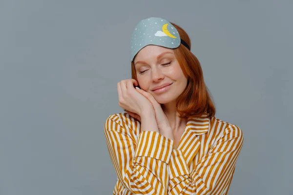 Pretty foxy freckled woman leans at palms, keeps eyes closed, wears sleepmask and stiped pajamas, dreams about something pleasant, stands indoor over grey wall, with copy space for promotion