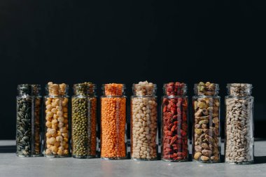 Glass bottles with sunflower seeds, mung beans, mush beans, lentils, pistachio, pumpkin seeds standing in one row against black background. Food storage