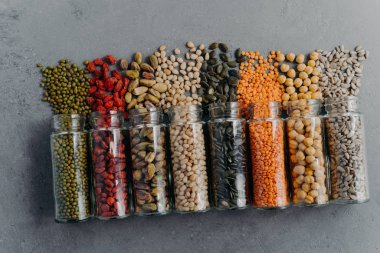 Indoor shot of organic grains, nuts, seeds in transparent glass jars spilled on grey surface. Food package storage. Healthy products for vegetarians. Zero waste products