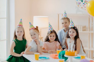 Çocuklar, kutlama ve doğum günü konsepti. Pozitif çocuklar partide eğleniyorlar, koni şapkaları takıyorlar, lezzetli pastalar yiyorlar, ilginç oyunlar oynuyorlar, keyifleri yerinde.
