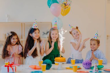 Çocuklar ve tatil konsepti. Beş arkadaşın pastada mutlu bir şekilde ışıltıyla görünmesine, doğum gününü kutlamasına, parti konisi şapkaları giymesine, balon tutmasına, mutlu ifadelere sahip olmasına sevindim.