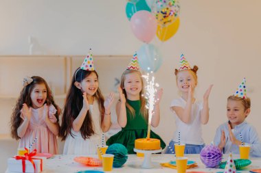 Coşkulu çocuklar el çırpar ve pasta üzerindeki büyük pırıltıya bakar, hediyeli şenlik masasının yanında durur, fincanlar ve tatil özellikleri, birlikte eğlenir, doğum gününü kutlar.