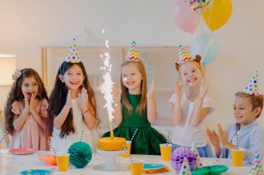 Tatil ve festival konsepti. Mutlu beş küçük çocuk el çırpıyor, pastadan çıkan ışıltıya bakıyor, arkadaşlarının doğum gününü kutluyor, büyük masanın yanında toplanıyor, balonlarla süslü bir odada duruyor.