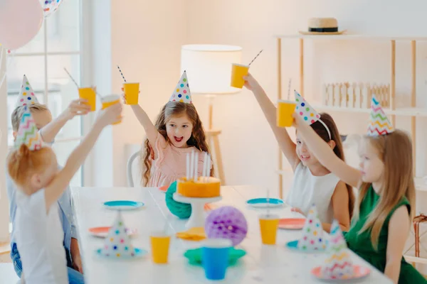 Des groupes d'enfants d'âge préscolaire joyeux célèbrent leur anniversaire ensemble, s'amusent, se réjouissent avec des tasses de boisson, portent des chapeaux de fête, mangent un délicieux gâteau, s'assoient à table dans une pièce spacieuse. Enfants et concept de parti — Photo