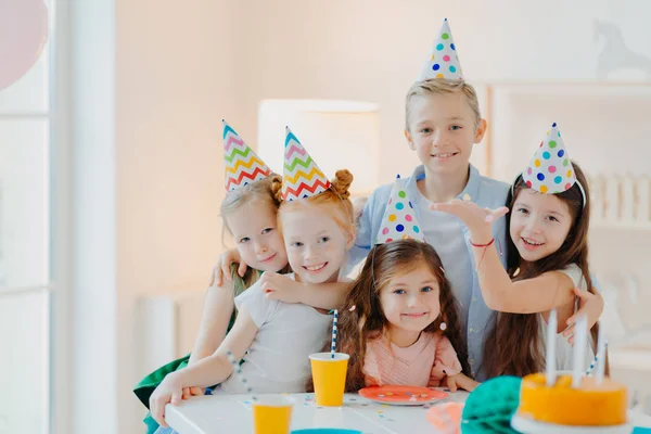 Foto interior de niños felices celebrar fiesta con confetti caída, llevar sombreros de la fiesta del cono, posar cerca de la mesa festiva con tartas, abrazarse y poner juntos. Niños cumpleaños —  Fotos de Stock