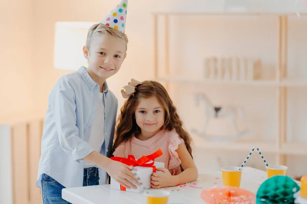 Menina pequena feliz e menino seguram caixa de presente com fita vermelha, preparar surpresa no aniversário, posar na mesa branca, usar chapéus de festa cone, têm expressões positivas. Conceito de infância — Fotografia de Stock