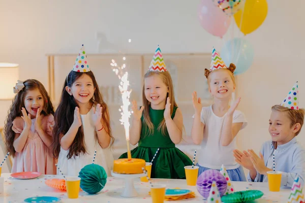 Amis sur la fête d'anniversaire, regarder avec plaisir au gâteau, se tenir debout près de la table de fête, porter des chapeaux cônes, taper des mains, jouer ensemble, poser dans une salle décorée. Concept d'enfance et de vacances — Photo