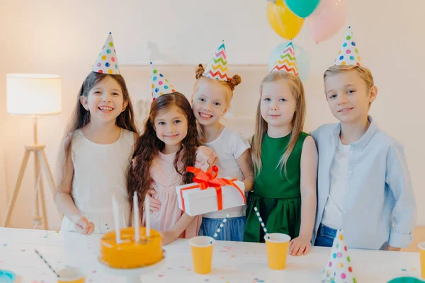 Glad birthday girl stands with presenet, happy friends come to congratulate her, wear party cone hats, stand near festive table with cake, smile joyfully, celebrate festive event. — Foto de Stock