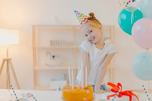 Malá zrzavá dívka nosí čepici a bílou košili, stojí u slavnostního stolu s dortem, fouká svíčky a přeje si během svých narozenin, pózuje v bílém pokoji s nafouknutými balónky, radostně se usmívá — Stock fotografie