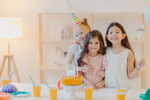 Horizontal shot of three happy friends embrace and have funny expressions, pose near festive table with cake indoor. Three girls celebrate birthday together, have fun, come to party — Stock Photo, Image