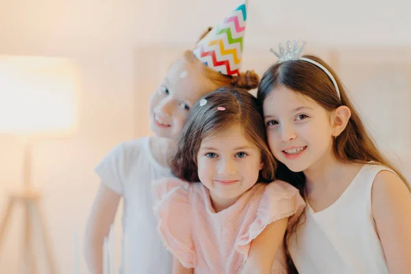 Retrato de tres amigos felices vestidos con ropa festiva, sombrero de fiesta, poner interior sobre fondo blanco, jugar juntos durante la celebración del cumpleaños. Las chicas guapas tienen buen humor, vienen en una ocasión especial. —  Fotos de Stock