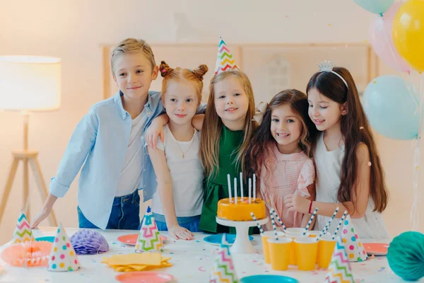 Los niños amistosos abrazan mientras se ponen cerca de la mesa festiva, soplan velas en el pastel, tienen humor de fiesta, celebran cumpleaños o ocasión especial, tienen expresiones alegres. Infancia, diversión y entretenimiento —  Fotos de Stock