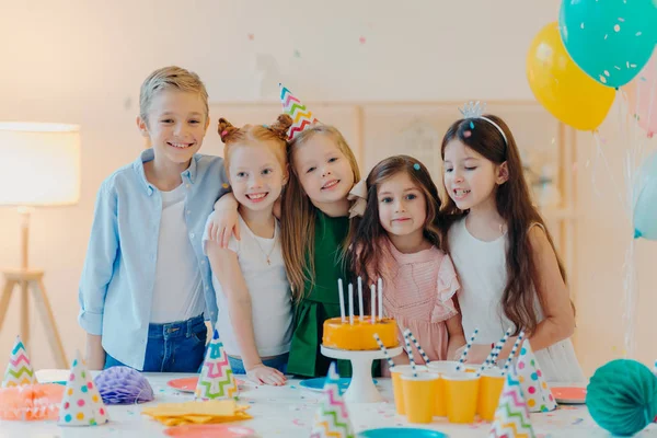 어린이들이 함께 모여 생일을 축하하고, 카메라를 들고 포즈를 취하고, 특별 한 행사를 준비하고, 케이크, 종이 컵, 파티 모자를 들고 테이블 가까이 서 있는 모습 이 수평으로 보인다 — 스톡 사진