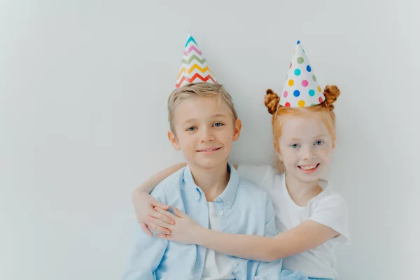 Una guapa chica roja de pelos abraza con amor a su hermano mayor, felicita con cumpleaños, lleva gorras de fiesta de cono, tiene buen humor en la fiesta, aislada sobre fondo blanco, tienen una relación amistosa. —  Fotos de Stock