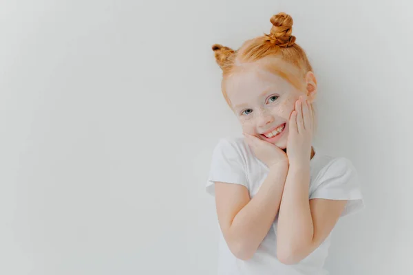 La chica guapa tímida tiene dos cabellos de pelo, toca mejillas suavemente, sonríe alegremente, tiene gafas, oye algo positivo de los padres, llora camiseta blanca, se pone interior, espacio en blanco dejado para su información. —  Fotos de Stock