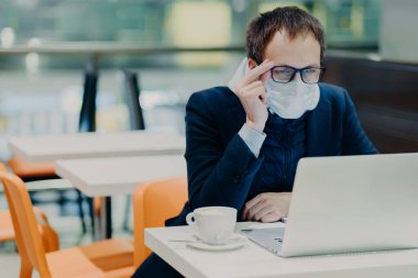 Tıbbi maskeli ve gözlüklü bir adam, modern dizüstü bilgisayarın önünde oturur, rahat kafeteryada poz verir, Coronavirus hakkında haberler okur, sağlıkla ilgilenir, kahve içer. Salgın Karantina