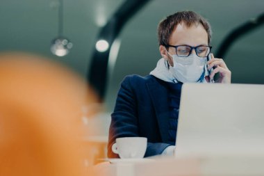 İşadamı kafeteryada uzaktan çalışıyor, dizüstü bilgisayar ekranında yoğunlaşıyor, telefonla konuşuyor, grip veya koronavirüs epidemisine karşı korunmak için yüzüne tıbbi maske takıyor. Akıllı çalışma