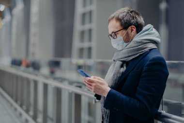 Erkek blogcuların yan fotoğrafları mesaj gönderiyor ve modern cep telefonundan takipçilerinin yorumlarını okuyor. Coronavirüs veya diğer bulaşıcı hastalıklardan korunmak için steril tıbbi maske takıyor.