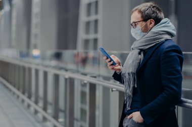 Ciddi Avrupalı adam akıllı telefona bakar, internetten haber okur, mesaj gönderir, virüsten korunur, tıbbi maske takar, koronavirüs korumasını önemser. Virüs salgını, Covid-19