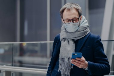 Adamın fotoğrafı telefonundan haberleri izliyor, koruyucu maske takıyor, hava kirlenmesinden ya da koronavirüsten koruyor, e-posta kutusunu kontrol ediyor, sıcak atkı takıyor. Covid-19. Salgın hastalık. Sağlık, sosyal medya