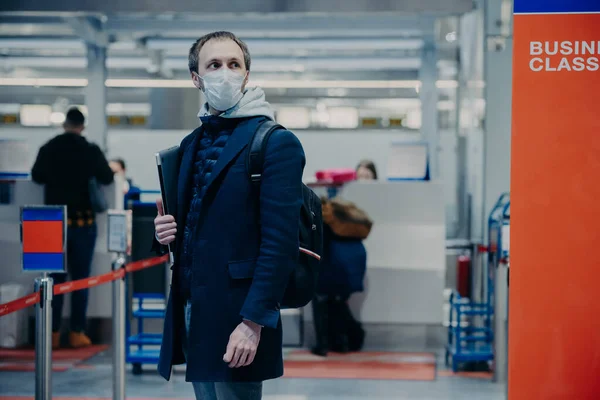 Man with luggage, wears medical protection mask, poses in airport terminal, travels by airplane during coronavirus outbreak, prevents disease, ready for fight and health checkpoint. Evacuated tourist