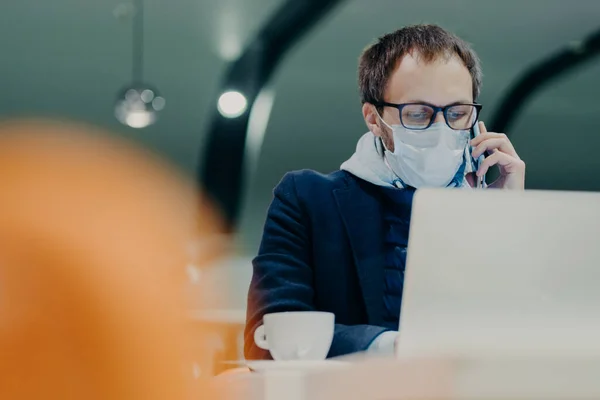 İşadamı kafeteryada uzaktan çalışıyor, dizüstü bilgisayar ekranında yoğunlaşıyor, telefonla konuşuyor, grip veya koronavirüs epidemisine karşı korunmak için yüzüne tıbbi maske takıyor. Akıllı çalışma