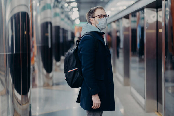 Preventive measures in public places. Sideways shot of man in medical mask stands on metro station, carries backpack, looks worried during epidemic situation. People, prevention, coronavirus