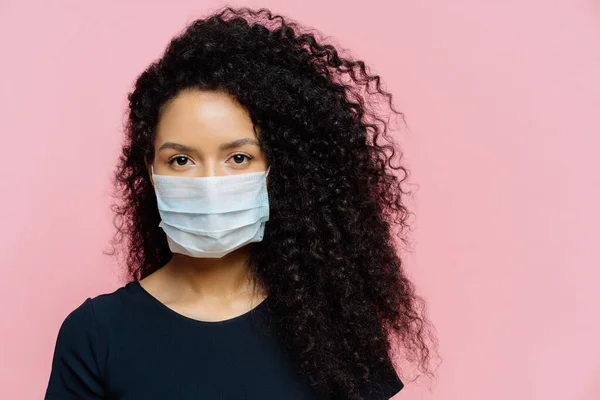 Ciddi koyu tenli genç bir kadın, evde kendini izole ediyor koruyucu tıbbi maske takıyor, evde karantinada, sıradan giyinmiş, pembe arka planda poz veriyor. Coronavirus hastalığı