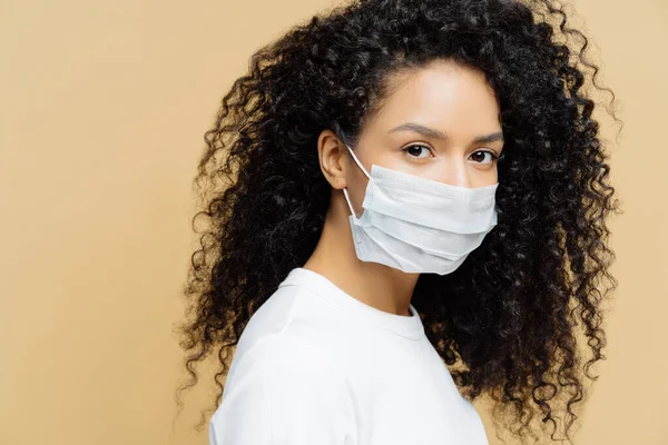 Koyu tenli, kıvırcık saçlı Afro-Amerikalı bir kadının fotoğrafı. Koronavirüs salgını sırasında koruyucu maske takıyor. Bej arka planda izole edilmiş. Hastalığı önle. Virüs, grip, sağlık hizmetleri.