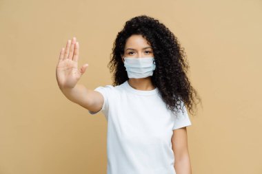 Serious dark skinned woman makes stop gesture, pulls palm towards camera, wears medical flu mask, asks stay at home not to spread coronavirus disease, prevents virus, dressed in white t shirt. clipart
