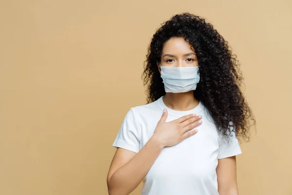 Ciddi Afro-Amerikalı kadın tıbbi maske takıyor, nefes almakta zorlanıyor, göğüs göğse baskı yapıyor, koronavirüs enfeksiyonu kapmış, bej kaplama bölgesinde izole edilmiş. Covid 19, sağlık hizmeti konsepti
