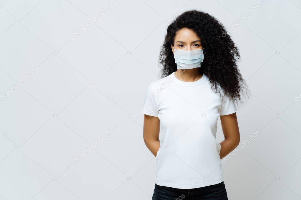 Half length shot of Afro American woman being on self isolation or quarantine, wears medical mask during coronavirus outbreak, listens news, tries not to spread virus, stays at home for long period