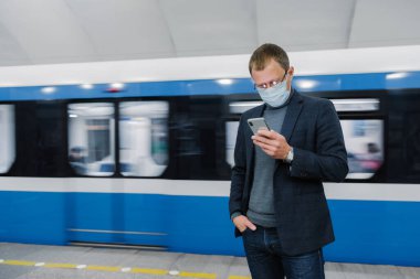 Erkek yolcu platformda yüz maskesi takıyor, treni bekliyor, yer altında gidip geliyor, akıllı telefon cihazına odaklanıyor, internetten haberleri okuyor. Virüs kamuya açık bir yerde. Coronavirüs salgını