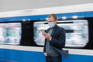 Gözlüklü ve koruyucu tıbbi maskeli bir adamın fotoğrafı, modern telefon kullanıyor, toplu taşıma araçlarıyla seyahat ediyor, platformda hareket halindeki trene karşı duruyor, yeni Covid-19 virüsü hakkında bilgi topluyor.